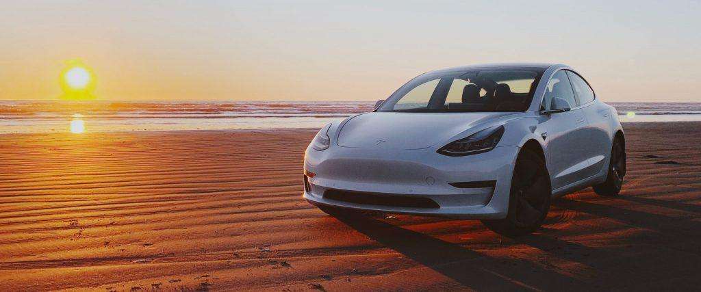 Tesla Model 3 parked on the beach with the sun setting in the background