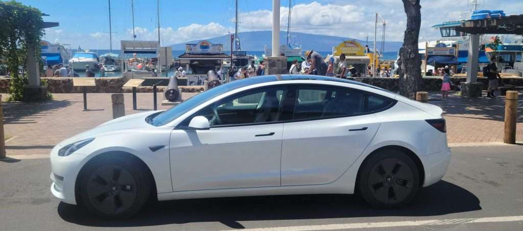 Tesla model 3 parked in front of a pier in Maui underneath a clear sky with some clouds