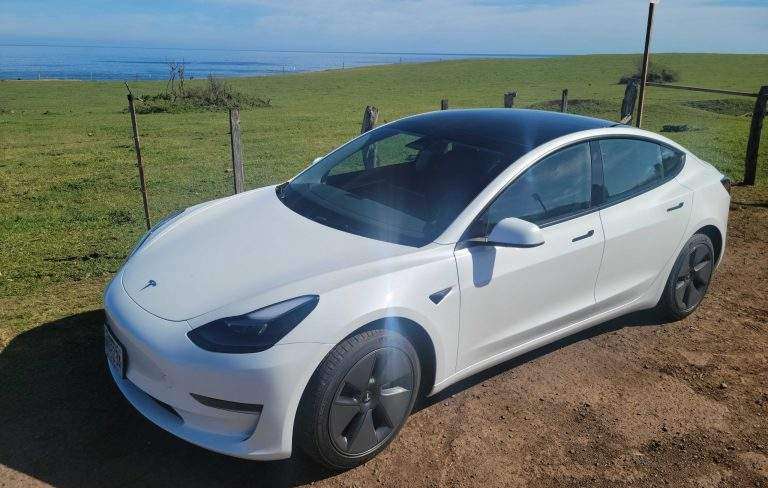 White Tesla Model 3 on red dirt with green grass, then blue ocean, then blue sky in the background