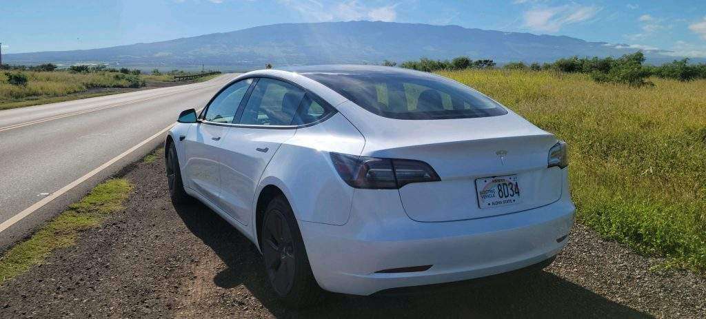 Tesla Model 3 on side of road and open skies, grass, and mountains in background