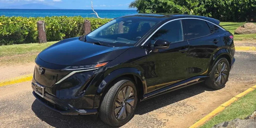2025 Nissan Ariya in Black Diamond finish, front-left angle with lush Maui landscape and ocean views.